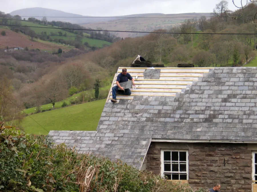 Roofer sitting on new battens while re-laying natural slates on an older stone cottage roof