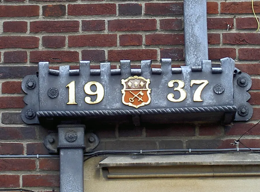 Ornamental lead rainwater head dated 1937 with a crest, fixed to a brick wall above a downpipe