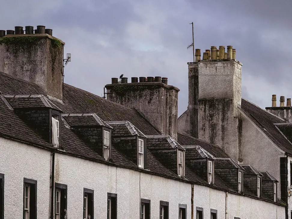 Row of rendered chimney stacks topped with clay pots above a slate-roofed terrace with dormer windows under a grey sky
